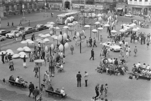Ein Schwarz-Weiß-Foto eines belebten Stadtplatzes mit Menschen, die gehen, auf Bänken sitzen und Lichtmasten, sowie Fahrzeugen auf der Straße und Gebäuden mit Fenstern und Namensschildern im Hintergrund.