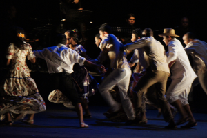 Eine Gruppe von Menschen tanzt im Vordergrund mit einer anderen Gruppe, Stühle und Mikrofone im Hintergrund, alles auf einem Boden.