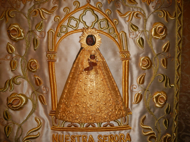 Goldene Stickerei mit einer serenen Frau in einem hochgeschlossenen, langärmligen Kleid mit einem Dutt, umgeben von detaillierten Texten und Designs in einem goldenen Rahmen.