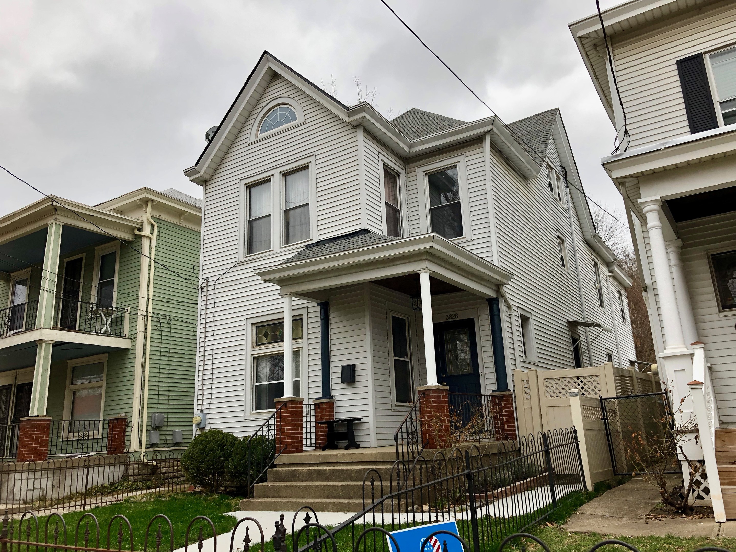 Reihe von Häusern mit Fenstern, Türen, Säulen, Geländern, Stufen und Dächern, umgeben von einem Metallzaun, Gras, Pflanzen, Bäumen und einem bewölkten Himmel, mit einem Mietschild unten, das die Adresse 5901 S Elmwood Ave, Chicago 60618 anzeigt.