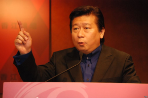 Ein Mann im Anzug und Krawatte hält eine Rede an einem Rednerpult mit Mikrofon und sieht wie ein Politologe mit ernster Miene aus.