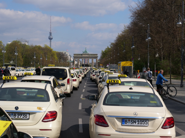 Eine lange Reihe von Taxis geparkt an einer belebten Straße in Berlin, Deutschland, mit Fahrradfahrern und Fußgängern auf dem Gehweg, flankiert von Bäumen und Laternenpfählen und Gebäuden, einem Bogen und einem Turm im Hintergrund unter einem bewölkten Himmel.