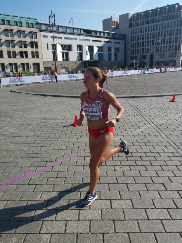 Eine Frau in einem Sportoutfit läuft durch eine Marathon-Strecke, die von Hütchen markiert ist und von Zuschauern umgeben ist.
