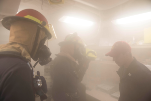 Eine Gruppe von Feuerwehrleuten in Helmen, mit Rauch aus dem Mund, einer hält eine Kamera, in einem Raum mit Lichtern und anderen Gegenständen, während einer Übung.