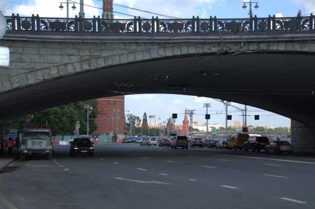 Eine Brücke spannt eine vielbefahrene Straße mit darunter fahrenden Fahrzeugen, während Fußgänger auf dem Gehweg links stehen, mit städtischen Elementen wie Ampeln, Bäumen, Gebäuden und Laternen im Hintergrund.