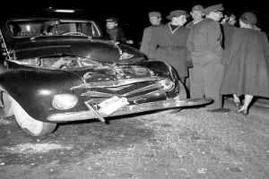 Eine schwarz-weisse Szene eines beschädigten Autos am Straßenrand, mit einer Gruppe von Menschen drumherum, die besorgt aussehen.