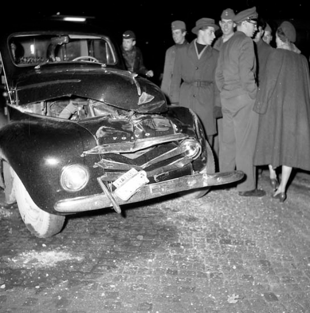 Eine schwarz-weisse Szene eines beschädigten Autos am Straßenrand, mit einer Gruppe von Menschen drumherum, die besorgt aussehen.