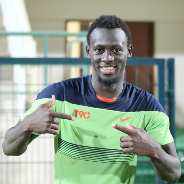 Ein Mann in einem grün-blauen Hemd, der lachend in die Kamera zeigt, mit einem unscharfen Geländer, einer Wand und einer Tür im Hintergrund.