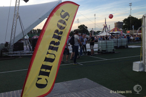 Eine lebhafte Karnevalszenen mit Zelten, einem zentralen Mast, Menschen, Bäumen, Strommasten, Straßenbahnen auf der Straße und einem Eisengestell links.