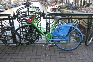Mehrere Fahrräder auf dem Boden geparkt mit einem Geländer dahinter, jenseits dessen sich ein Gewässer mit Booten befindet; Autos und Gebäude säumen beide Seiten des Gewässers, und es gibt Stühle und Tische in der oberen rechten Ecke.