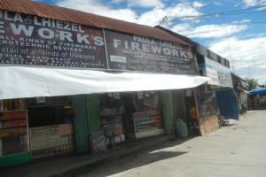Ein Geschäft mit einer 'Feuerwerk' -Schilder, umgeben von verschiedenen Gegenständen und Geschäften, mit Bäumen, Polen, Drähten und einem bewölkten Himmel im Hintergrund.