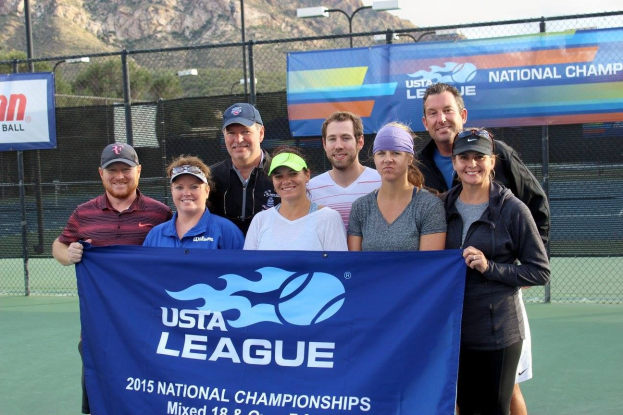 Gruppe von Menschen auf einem Tennisplatz mit einem "2015 USTA National Championships"-Banner, einem Zaun, Bannern, Laternenpf├Âhlen, Büschen, Bergen und einem klaren blauen Himmel im Hintergrund.