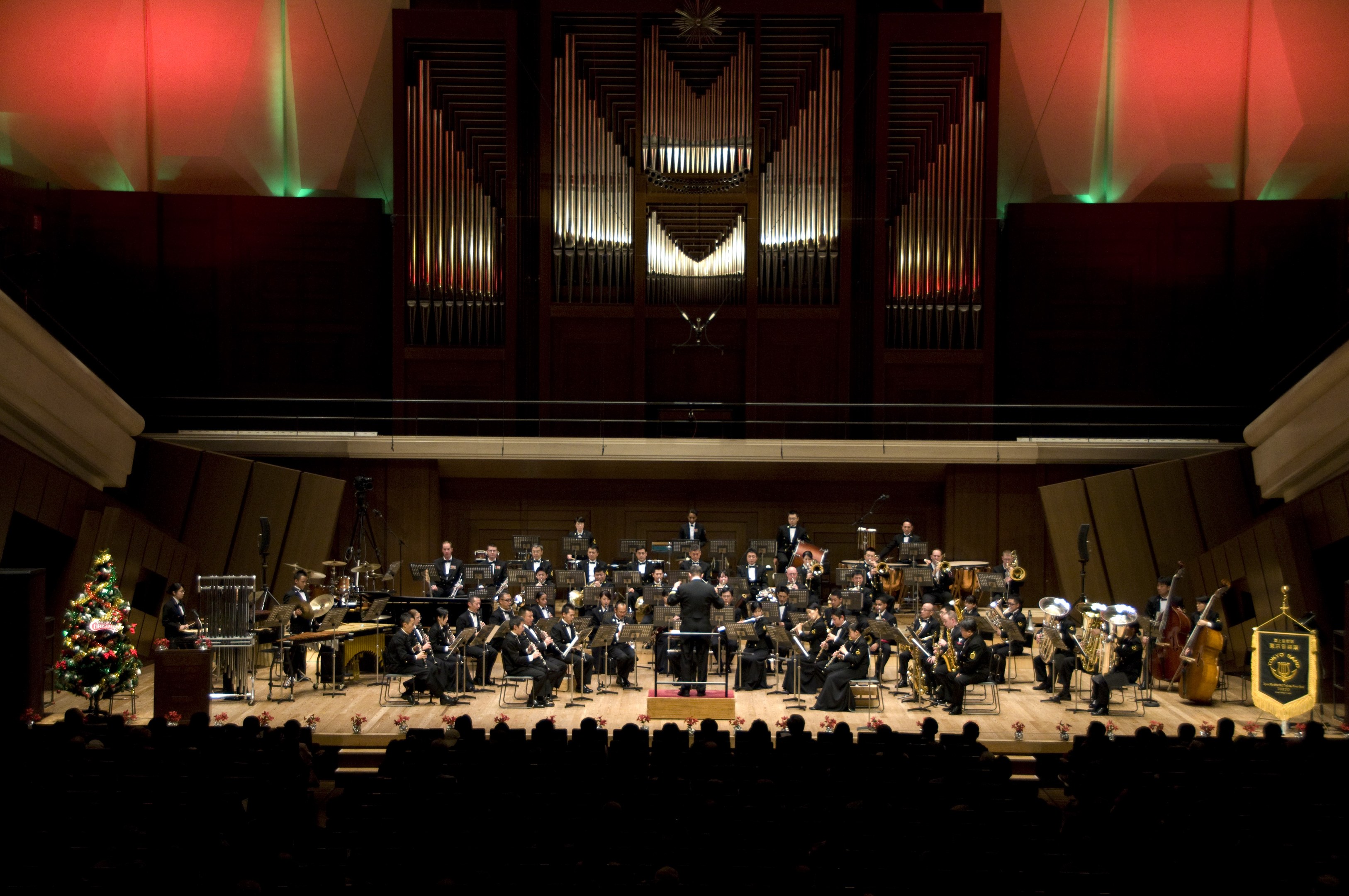 Großes Orchester auf der Bühne mit Weihnachtsbaum im Hintergrund, Musiker auf Stühlen sitzend, Dirigent mit Mikrofon auf einem Podium und Texttafeln rechts.