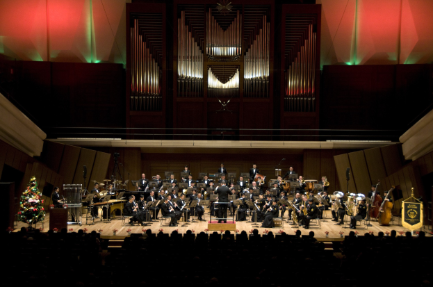 Großes Orchester auf der Bühne mit Weihnachtsbaum im Hintergrund, Musiker auf Stühlen sitzend, Dirigent mit Mikrofon auf einem Podium und Texttafeln rechts.