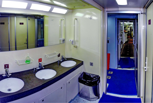 Badezimmer mit zwei Waschbecken, Wasserhähnen, einem Mülleimer, Schränken, einem Wandspiegel, Deckenleuchten und Menschen im Hintergrund.