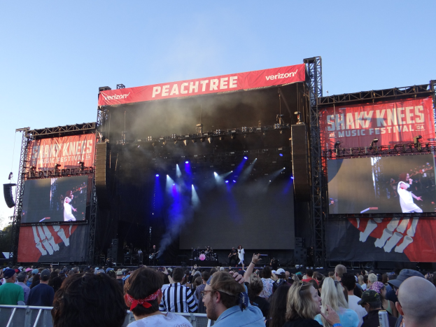 Eine große Menschenmenge steht vor einer hell beleuchteten Bühne auf dem Shaky Knees Music Festival, auf der Musiker unter Bannern mit Text spielen, mit einem Geländer im Vordergrund und einem klaren blauen Himmel im Hintergrund.