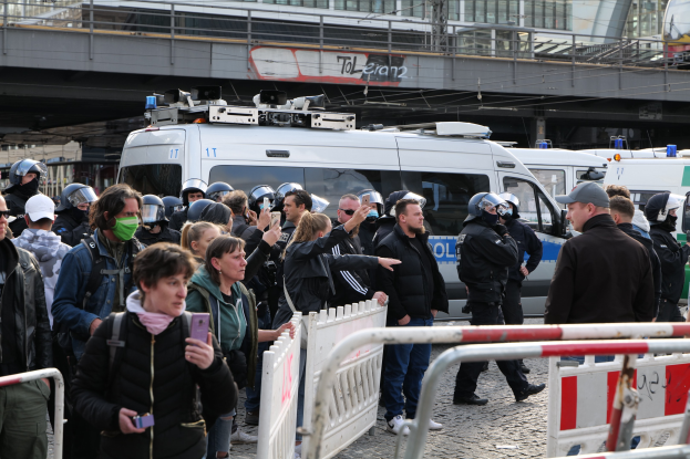 Eine Gruppe von Menschen steht hinter Barrikaden vor Polizeifahrzeugen während einer Demonstration in Berlin, einige tragen Helme und halten Telefone, mit einer Brücke und Gebäuden im Hintergrund.