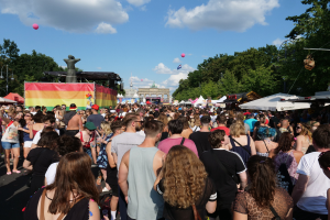 Eine gro├če Menge geht eine Stra├če entlang, die mit Zelten, B├Ąumen, Pfosten, Lichtern und einer Statue ges├Ąumt ist, w├Ąhrend des Berliner Pride-Festivals.