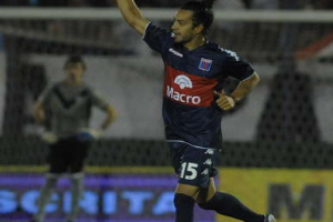 Ein Fußballspieler feiert nach einem Tor auf dem Boden, mit einer jubelnden Menge und einer Person im Hintergrund und einer Tafel mit Text dahinter, alles vor einem unscharfen, energetischen Hintergrund.