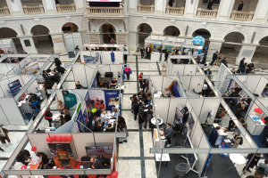 Eine große Gruppe von Menschen versammelt sich in einem Raum voller Stände, Banner, Tische, Stühle, Luftballons und einer Gebäudefront mit Bögen, Säulen, Geländern und einem Bildschirm im Hintergrund.