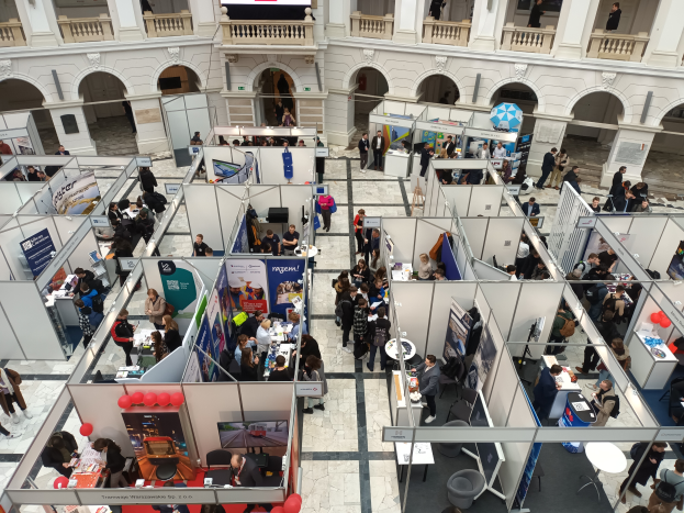 Eine große Gruppe von Menschen versammelt sich in einem Raum voller Stände, Banner, Tische, Stühle, Luftballons und einer Gebäudefront mit Bögen, Säulen, Geländern und einem Bildschirm im Hintergrund.