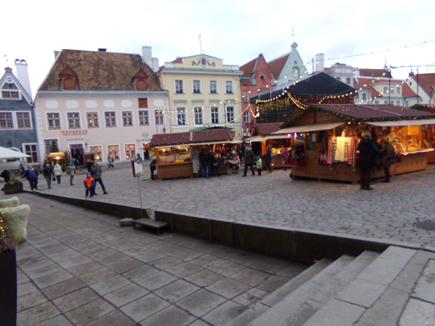 Ein lebhafter Weihnachtsmarkt in Tallinn, Estland mit Menschen um geschmückte Stände, festlicher Beleuchtung, Gebäuden im Hintergrund, bewölktem Himmel, Treppen und Topfpflanzen.