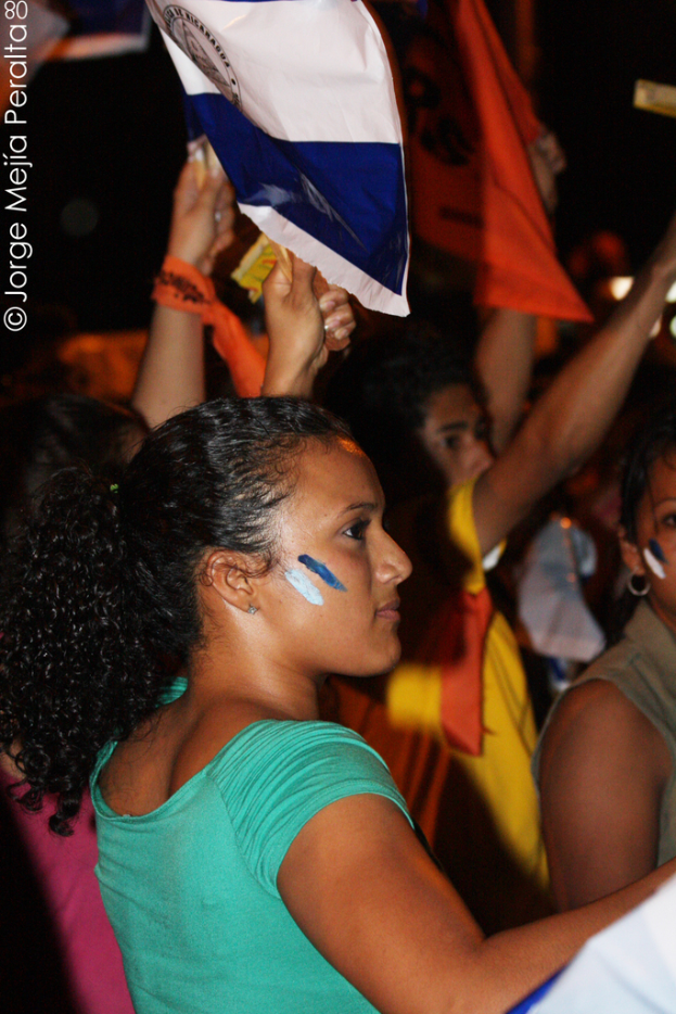 Eine Frau in einem grünen T-Shirt steht auf einer Demonstration und hält ein weißes Schild in der Hand, mit vielen anderen Menschen hinter ihr.