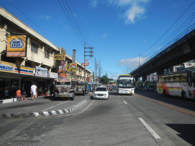 Eine belebte Stra{\u}enlandschaft mit Autos, Bussen, Fussgängern, Gebäuden, Strommasten, einer Brücke, Bäumen und einem bewölkten Himmel.