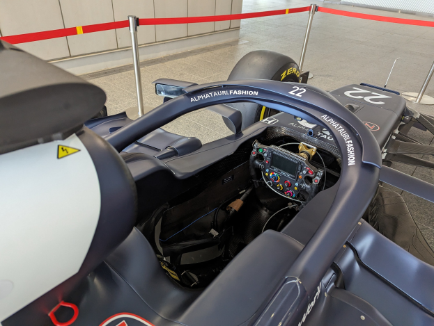 Cockpit eines Formel E-Autos in einer Ausstellungshalle mit einem Lenkrad im Vordergrund, einer Wand im Hintergrund und um das Auto herum gespannten Pfosten.
