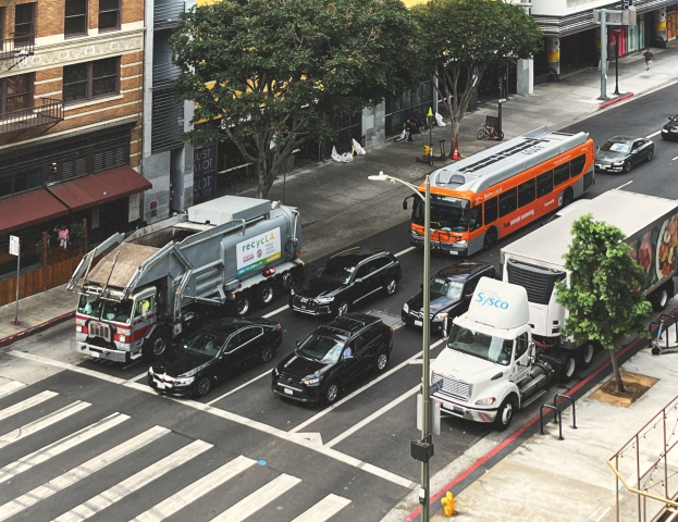 Eine belebte Stadtstraße mit Autos, Bussen und Lastwagen, gesäumt von Bäumen, Laternenmasten und Gebäuden, mit Geländern, Treppen, Fußgängern auf dem Gehweg und Texttafeln.