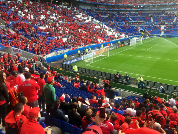 Eine große Menschenmenge in einem Stadion schaut einem Fußballspiel zu, mit Sitzplätzen und Stehplätzen, Bannern, einem Torpfosten und Deckenleuchten unter einer Dachkonstruktion.