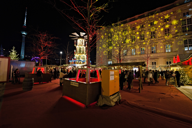 Ein geschäftiges Weihnachtsmarkt in Berlin, Deutschland mit Menschen um geschmückte Stände, festliche Lichter, Tannenbäume, Gebäude, Laternenmäste und einen Turm unter einem dunklen Himmel.