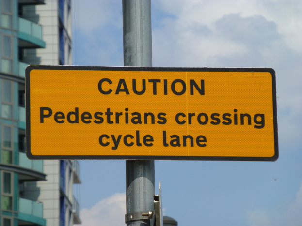 Gelbes Warnschild an einem Pfahl vor einem Gebäude, auf dem steht: "Fußgänger überqueren Fahrradspur" vor einem klaren blauen Himmel.