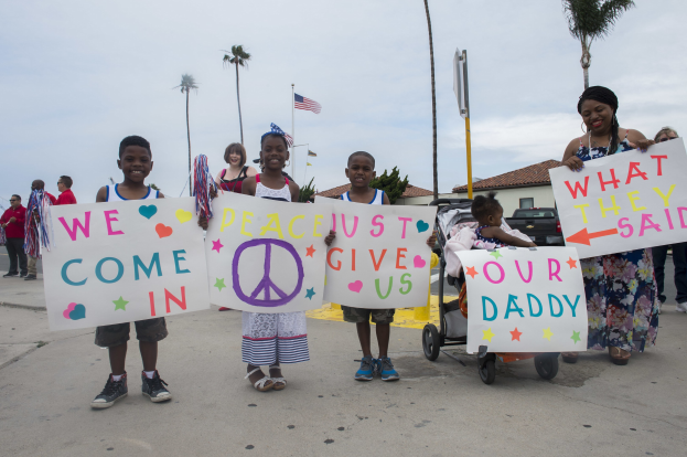 Eine Gruppe von Kindern hält Schilder und steht auf einer Straße vor einem Gebäude, während eine Frau einen Kinderwagen mit einem Baby schiebt; auf einem Schild steht "Wir kommen in Frieden, gebt uns unseren Daddy."