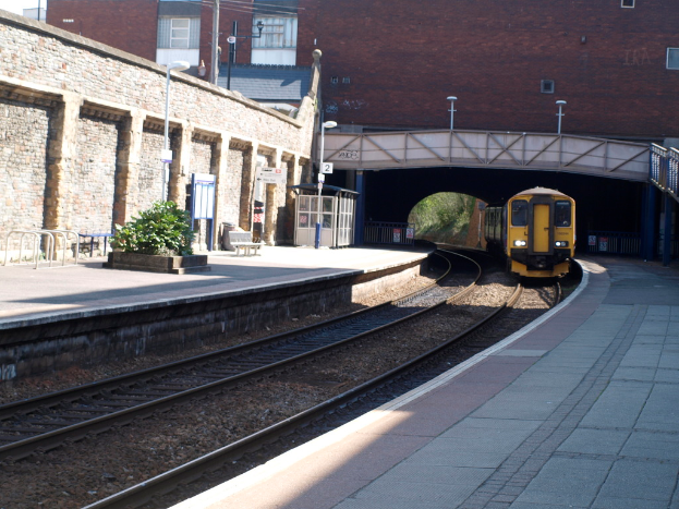 Ein Zug fährt auf den Schienen mit Plattformen auf beiden Seiten, eine Hauswand links, Straßenlaternen, Pflanzen, Bänke, eine Hütte, eine Brücke über die Plattformen und Gebäude und Masten im Hintergrund.