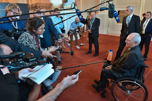 Ein Mann im Rollstuhl in Anzug und Krawatte ist von Reportern mit Kameras und Mikrofonen umgeben, mit einem Banner und einer Wand im Hintergrund.