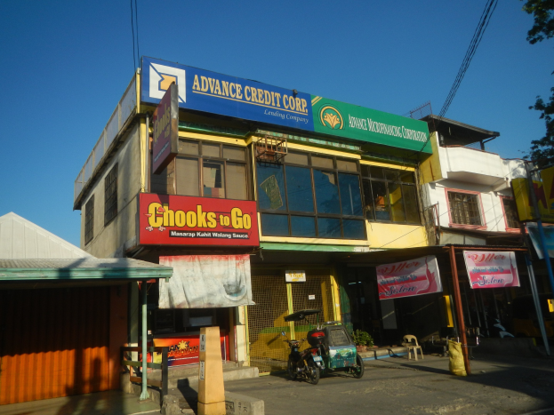 Ein Gebäude mit einer Tafel, auf der 'Advance Credit Corp' steht, an einer Straße mit Fahrzeugen, Werbetafeln, Strommasten, Kabeln, Bäumen und einem klaren blauen Himmel.