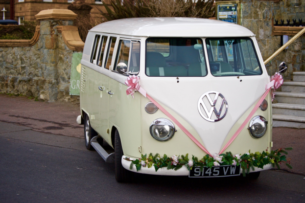 Ein weißer und pinkfarbener VW-Camper mit floralen Verzierungen, der vor einer Steinmauer geparkt ist, mit einer Treppe auf der rechten Seite und einem Gebäude im Hintergrund.