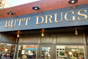 Außenansicht eines Gebäudes mit einer beleuchteten Schilder 'butt drugs', mit Glaswänden und -türen, mit Fahrzeugen, Gebäuden, Bäumen und Himmel durch das Glas zu sehen.