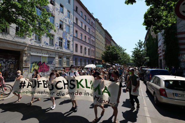 Eine Gruppe von Menschen, die mit einem Spruchband die Straße entlanggehen, mit parkenden Autos auf der rechten Seite, einem Fahrradfahrer auf der linken Seite, Gebäuden und Bäumen im Hintergrund und einer Berliner Protestszene.