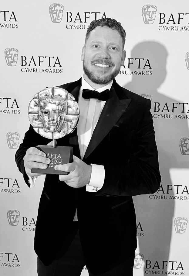 Ein Mann in einem Smoking, der eine BAFTA-Trophäe hält und lächelt, mit einem Hintergrund, der "BAFTA Awards 2018" anzeigt.
