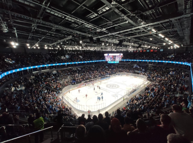 Hockey-Spiel in einer großen Arena mit Zuschauern auf den Rängen und Personal am Boden, mit Geländern, Deckenbeleuchtung und einem Bildschirm oben, identifiziert als O2 Arena in London.