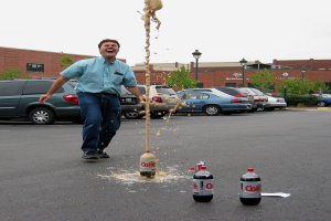 Ein Mann in einem blauen Hemd und Jeans steht lachend auf einer Straße außerhalb der Stadt, mit drei leeren Flaschen in der Nähe, acht geparkten Autos, Bäumen, einer Straßenlaterne und einem roten Backsteingebäude im Hintergrund.