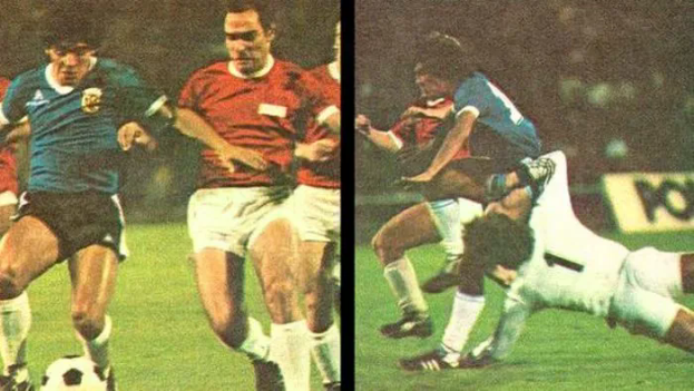 Eine Gruppe von Männern, die auf einem Feld Fußball spielen, wobei einer den Ball tritt und andere versuchen, die Kontrolle zu erlangen, vor einer Tafel in einem Stadion.