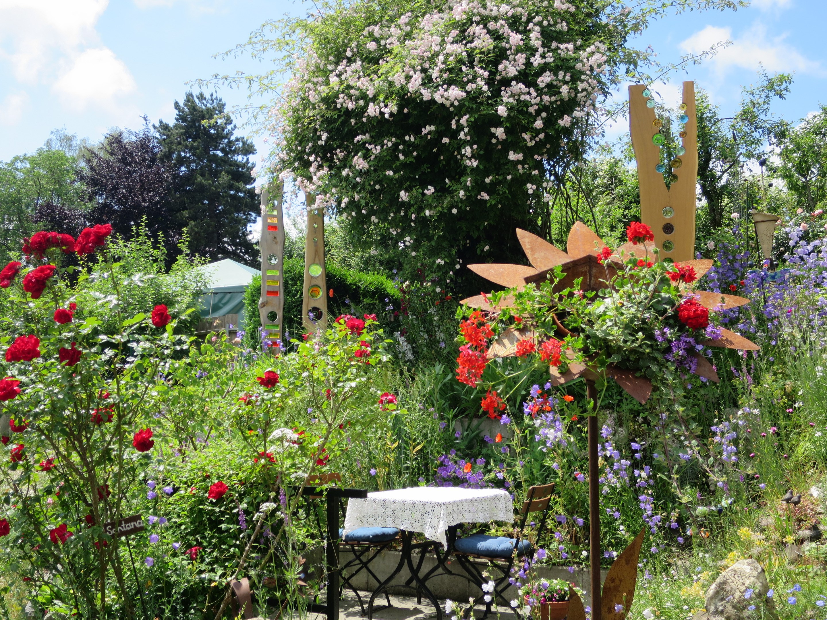 Ein Garten mit bunten Blumen und Pflanzen, der einen Tisch mit einer Decke und Stühlen in der Mitte zeigt, umgeben von Bäumen unter einem bewölkten Himmel auf dem Chelsea Flower Show.