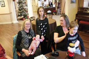 Ein Raum mit einer Wand, die ein Fotoframe, einen Weihnachtsbaum, eine Lampe, Pflanzen und Blumen zeigt. Es gibt Treppen, einige Menschen, die auf Stühlen und Tischen sitzen, mit einem Spielzeug und einer Kamera auf einem der Tische.