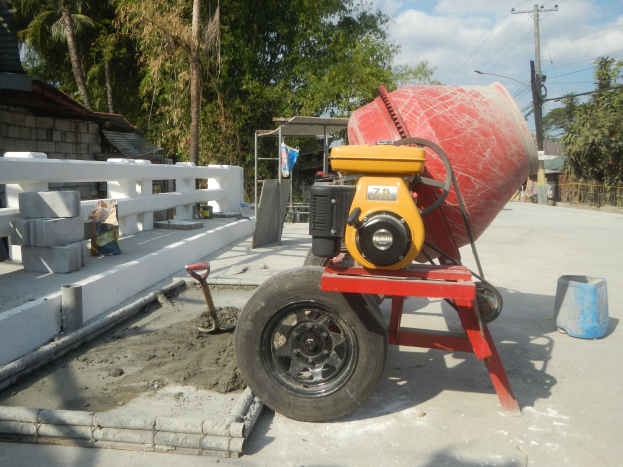 Betongrührer auf einem Zementlaster auf einer Straße mit Betonblöcken, einem Eimer, einem Handwagen, einem Zaun, einem Haus, Bäumen, Strommasten mit Drähten und einem bewölkten Himmel.