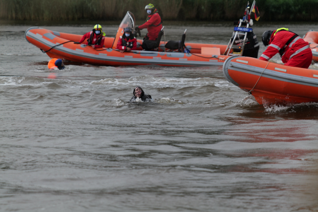 Gruppe von Menschen in einem aufblasbaren Boot auf einem Fluss, mit zwei Personen im Wasser im Vordergrund und Vegetation im Hintergrund, alle tragen Schwimmwesten und Helme während einer Rettungsaktion.