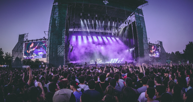 Eine große Menge Menschen bei einem Musikfestival schaut auf eine hell erleuchtete Bühne mit Instrumenten und Bildschirmen, die vor einem Gebäude, Bäumen und einem klaren blauen Himmel steht.