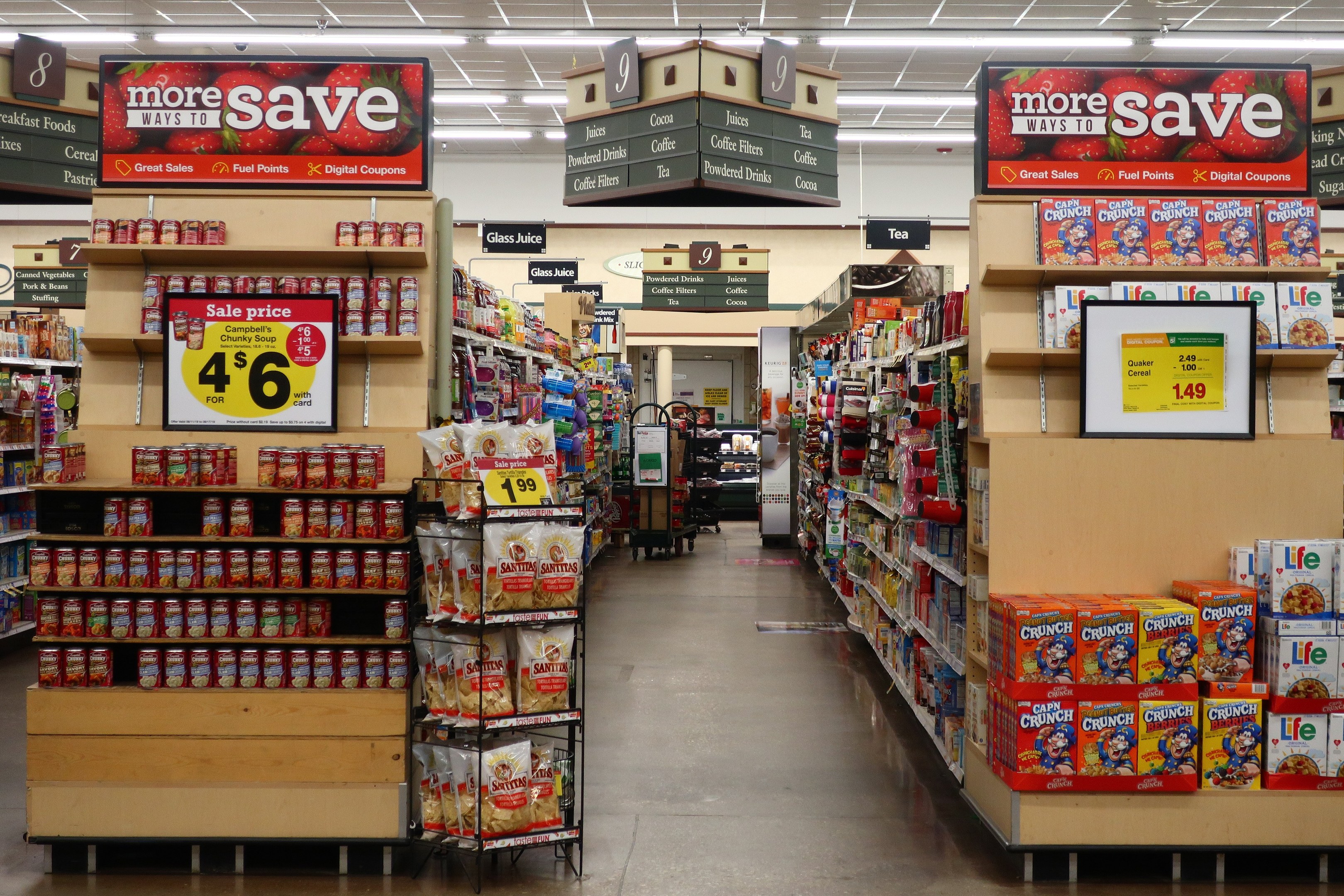 Ein Kiosk-Innenraum mit Regalen und Boden voller verschiedener Lebensmittel, beleuchtet von Deckenlampen und mit Informationsschildern mit Text und Zahlen.